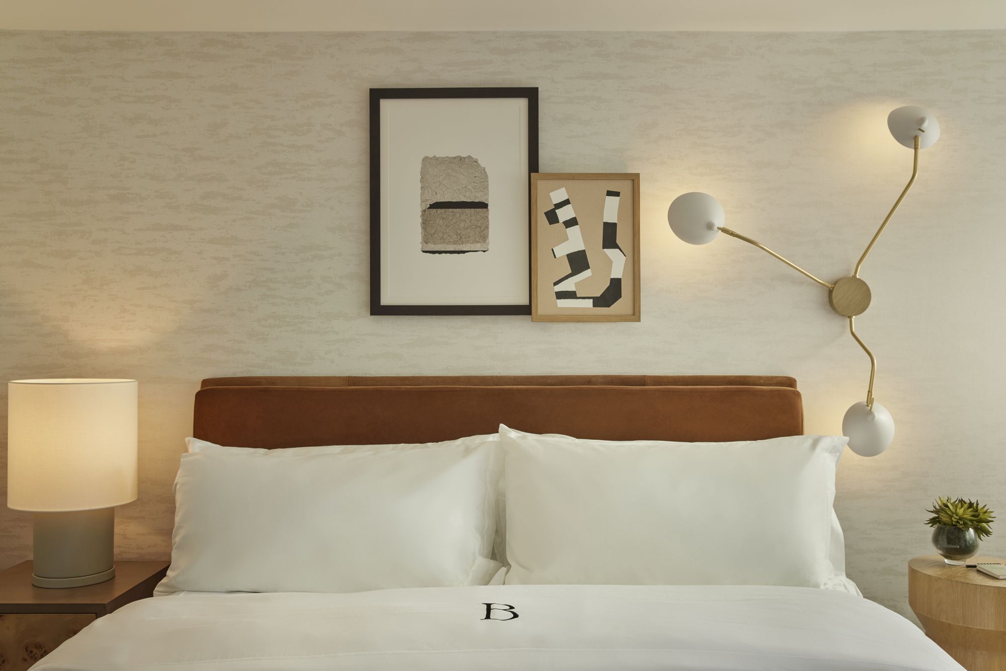 A neatly made bed with white linens and modern decor, including abstract art on the wall, a unique wall lamp, and a bedside table.