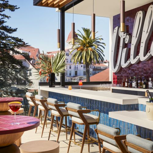 The image shows a stylish outdoor bar with a row of wooden chairs, modern tables, palm trees, and a scenic view in the background.