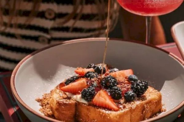 A plate of toast with strawberries and blackberries, topped with syrup, next to a pink cocktail with a flower garnish.