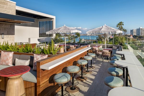 The image shows a rooftop terrace with tables, chairs, and umbrellas, overlooking a scenic view of the ocean and distant buildings.