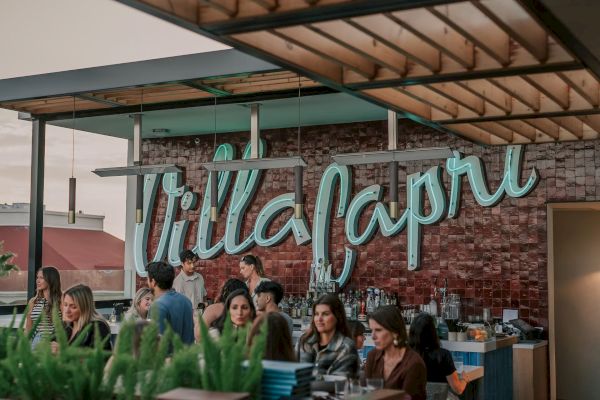 A group of people are socializing at an outdoor venue with a sign that reads "Villa Capri," surrounded by a modern and stylish decor.