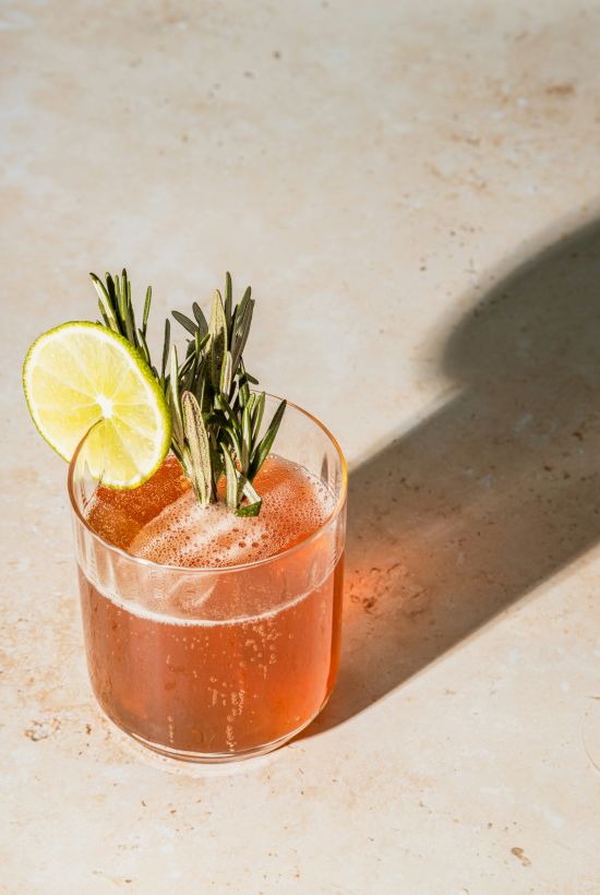 A cocktail with a lime slice, rosemary garnish, and ice cube in a clear glass, casting a shadow on a light surface.