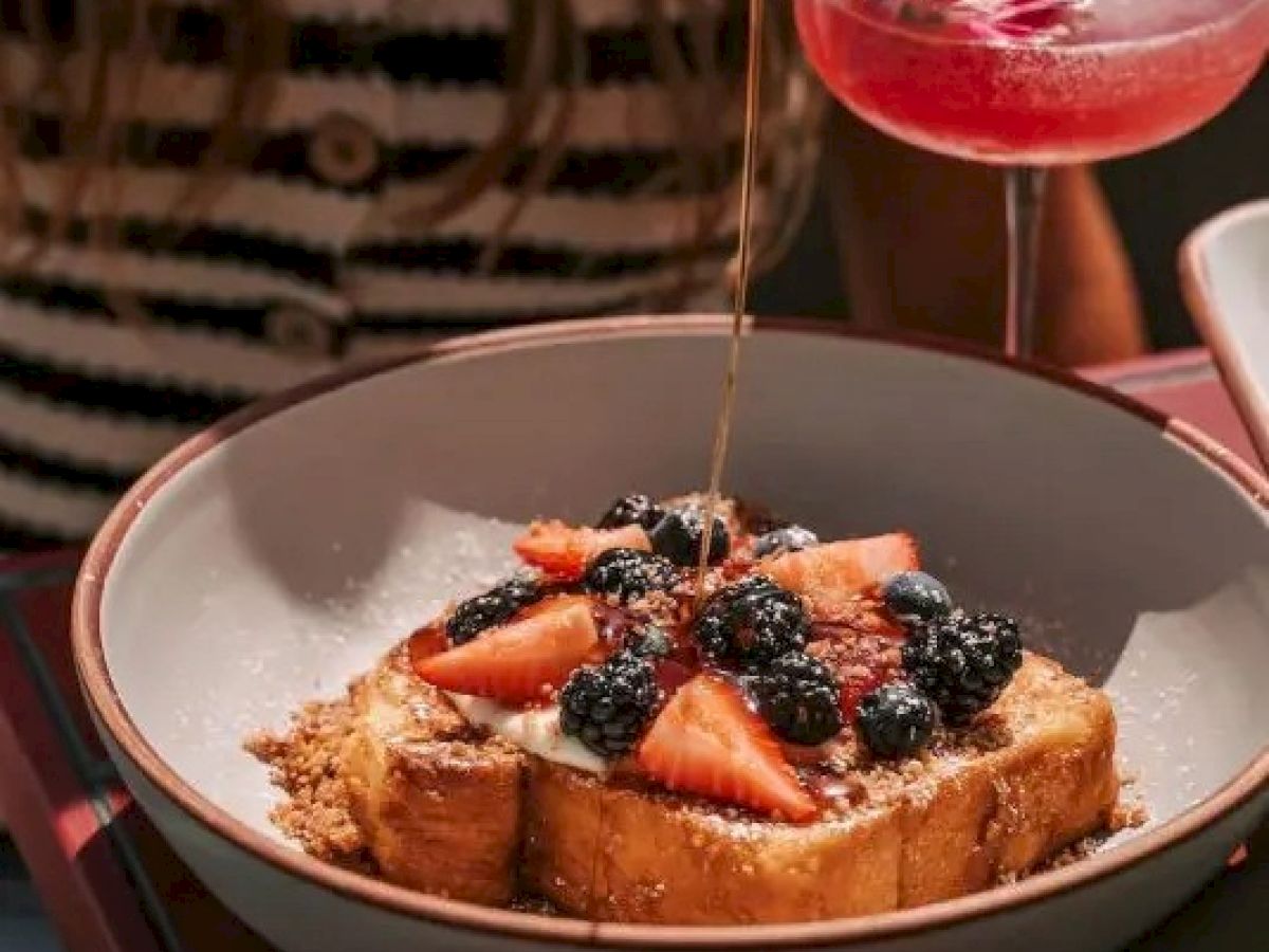 A bowl of French toast topped with strawberries, blackberries, and syrup, beside a cocktail with a pink flower garnish.