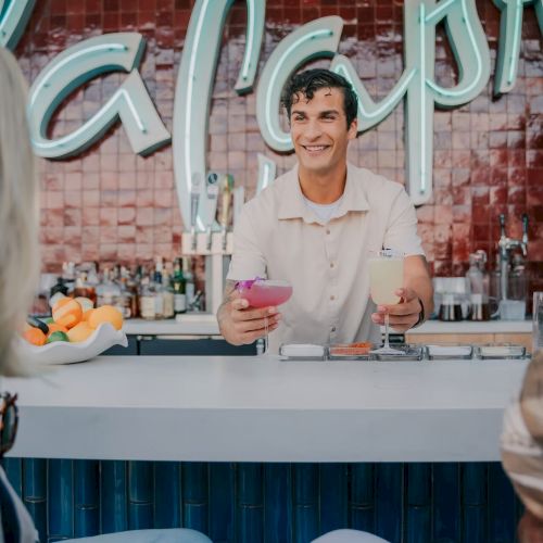 A bartender is serving drinks at a bar. A neon sign and bottles are in the background, with customers seated at the counter.