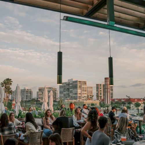 A bustling outdoor restaurant with people dining, surrounded by tall buildings and palm trees under a cloudy sky.
