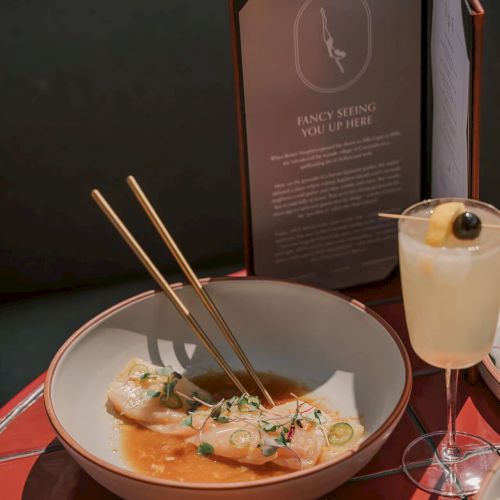 A bowl of gourmet food with chopsticks, a beverage, and an open menu on a table.