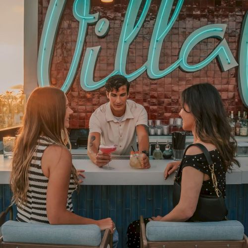 Two women sit at a bar while a bartender serves drinks. A neon sign reading "Villa" is visible in the background, adding ambiance.