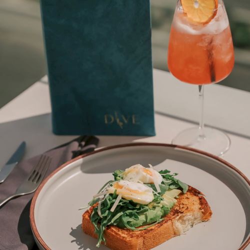 A plate with toast, avocado, eggs, and greens, alongside a pink drink with a slice of orange, placed in a scenic outdoor setting.
