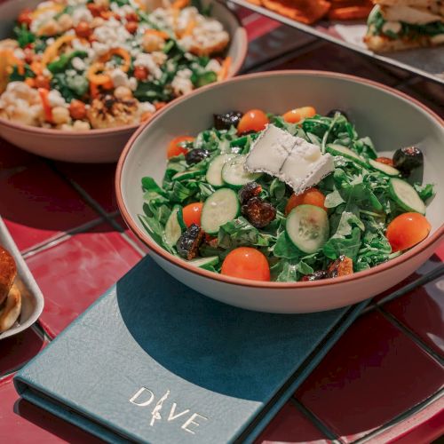 A table with a salad featuring greens, cucumbers, tomatoes, cheese, and a menu labeled "DIVE." Plates of food are visible in the background.