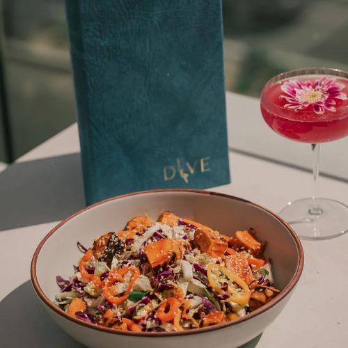 A colorful salad in a bowl, a pink cocktail with a flower garnish, and a blue menu are on a table.