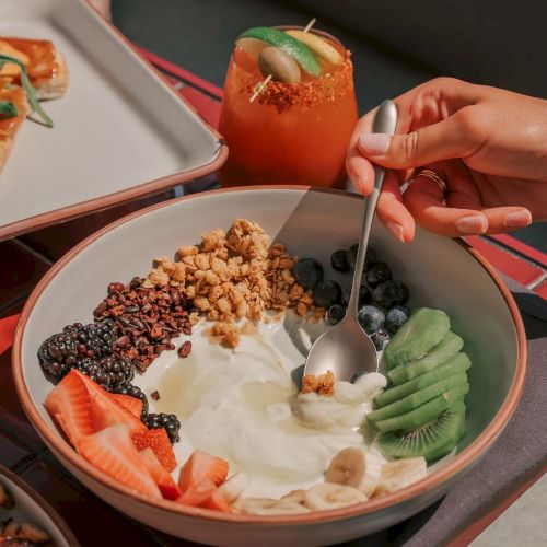 A bowl with yogurt, fruits, nuts, and a person's hand holding a spoon. A drink with lime is in the background on a tray.