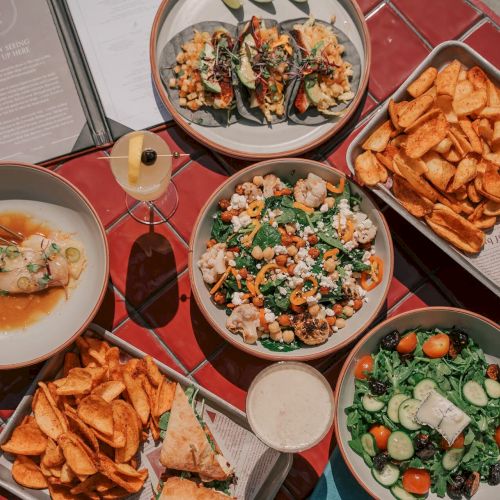 An array of dishes including tacos, a salad, sliders, and chips on a round red table with a drink and menu.