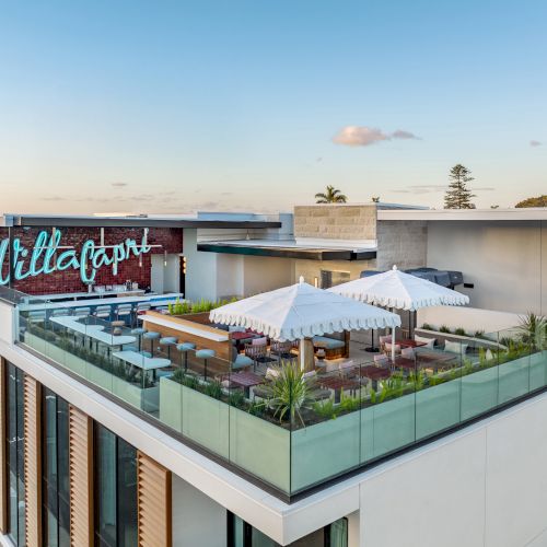 A rooftop restaurant with outdoor seating, umbrellas, and decorative plants. A neon sign reads "Villa Capri." The view includes a clear sky.