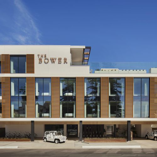 A modern three-story building named "The Bower," featuring large windows, sleek design elements, and parked white vehicles in front.