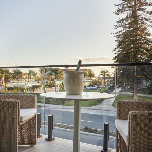 A small outdoor table with a champagne bottle in a cooler, surrounded by chairs, overlooking a road and trees under a clear sky.