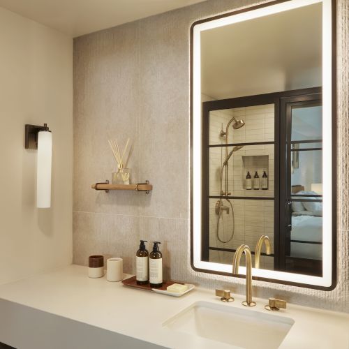 A modern bathroom with a lit mirror, gold faucet, toiletries, and a wineglass design on the counter; also visible is a shower area.