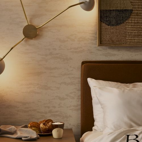 A cozy bedroom corner with a lit wall lamp, framed artwork, nightstand with items, and a neatly made bed with white linens.