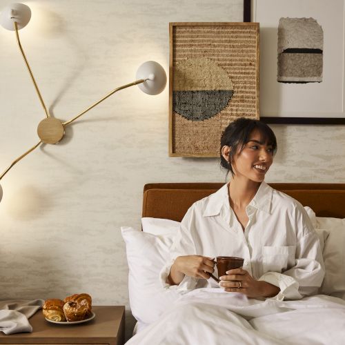A person is sitting in bed with a cup, near a bedside table holding pastries. The wall features modern artwork and a unique lamp design.