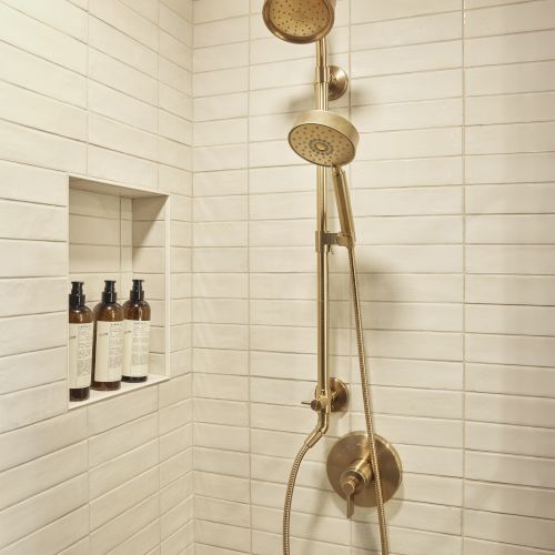 A shower with a gold fixture, dual showerheads, and three bottles on a shelf in a white-tiled bathroom.