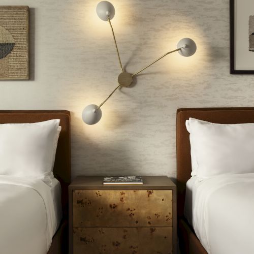 A modern bedroom with two beds separated by a wooden nightstand, featuring a unique wall-mounted light fixture.