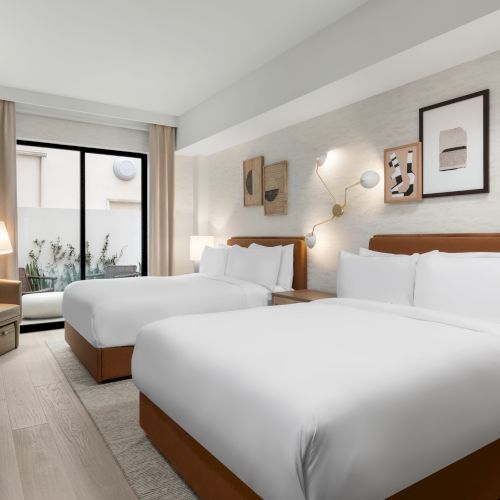 The image shows a modern hotel room with two beds, stylish decor, a window with curtains, and a cozy seating area near the window.