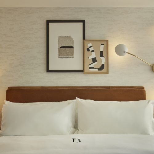 A modern bedroom with a neatly made bed, abstract wall art, unique wall light, and bedside tables with a lamp and a small plant.