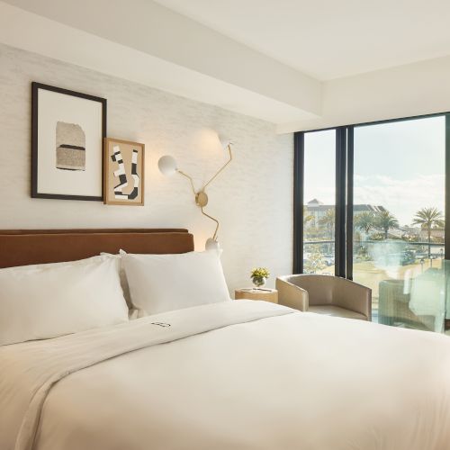 A modern hotel room with a neatly made bed, artwork, large windows, a chair, and a small table overlooking an outdoor balcony or view.