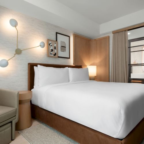 A modern hotel room with a large bed, stylish wall art, a lamp, and a chair. A sliding glass door leads to another area.