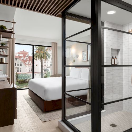 The image shows a modern hotel room with a glass shower, a neatly made bed, a large window with a view, and stylish wood accents.