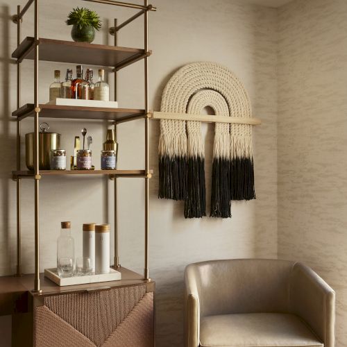 A cozy corner features a stylish shelf with bottles, a wall hanging with a semicircular design, and a cream armchair with a wooden side table.