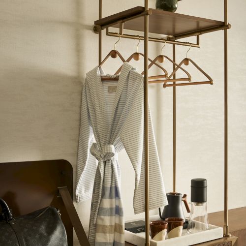 A clothing rack with a bathrobe, hangers, a plant, luggage, and a coffee setup on a wooden shelf in a cozy room setting.