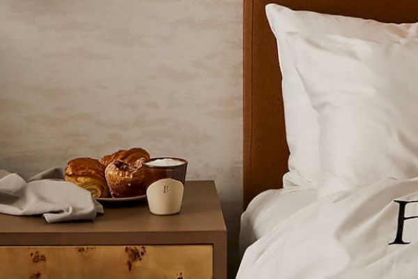 A nightstand with pastries, a cup, and a lamp next to a bed with white linens and a brown headboard.