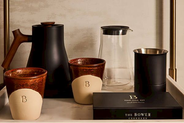 A tray holds a black pitcher, clear carafe, ceramic mugs, a cup, and a black box labeled "The Bower" in elegant packaging.