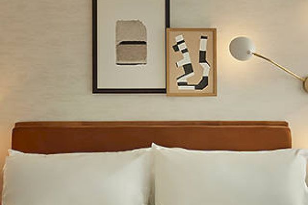 A neatly made bed with white pillows, two abstract art pieces above, and a wall-mounted lamp beside it.