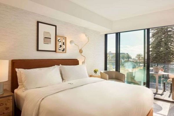 A cozy hotel room features a large bed, art on the wall, modern lighting, and a window with a view of trees and the outdoors.