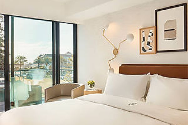 A modern hotel room with a neatly made bed, wall art, a window view of a pool and palm trees, and minimalistic decor.