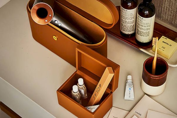 A Dyson hairdryer in a brown case, toiletries including bottles and tubes, a toothbrush in a holder, and Aesop products on a bathroom counter.