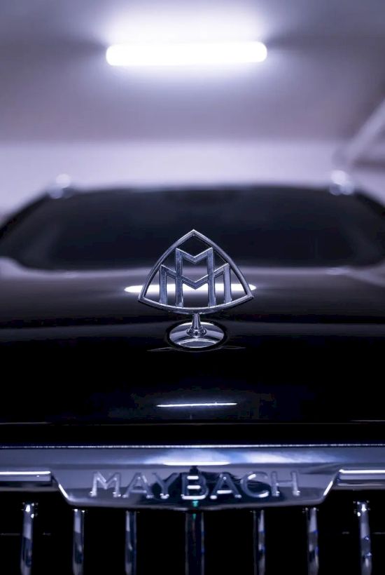 The image shows a close-up of a Maybach car, highlighting the emblem and the grill with the Maybach logo in a dimly lit setting.