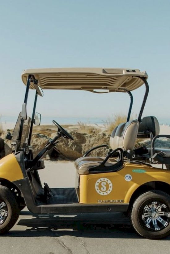 A yellow golf cart with a logo is parked on a street near the beach and rocks, under a clear blue sky.