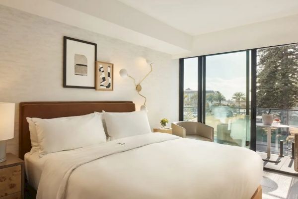 A cozy hotel room with a large bed, modern wall art, soft lighting, and a balcony offering a scenic view of trees and water.